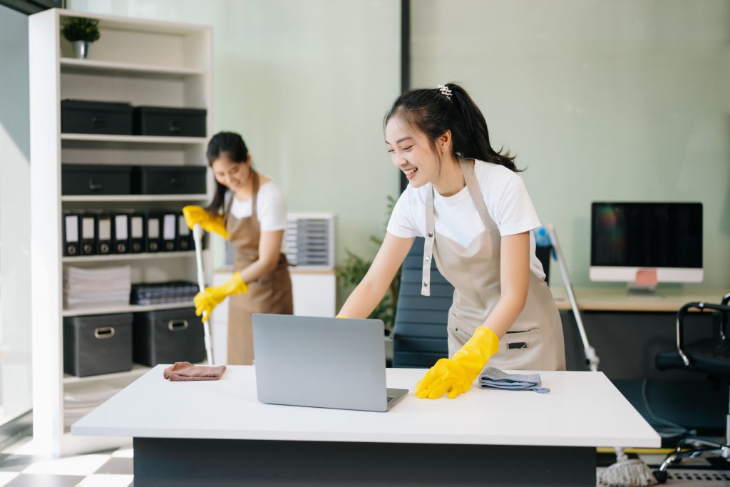 Two women wearing white shirts and beige aprons cleaning a modern office space with a wipe and mop.